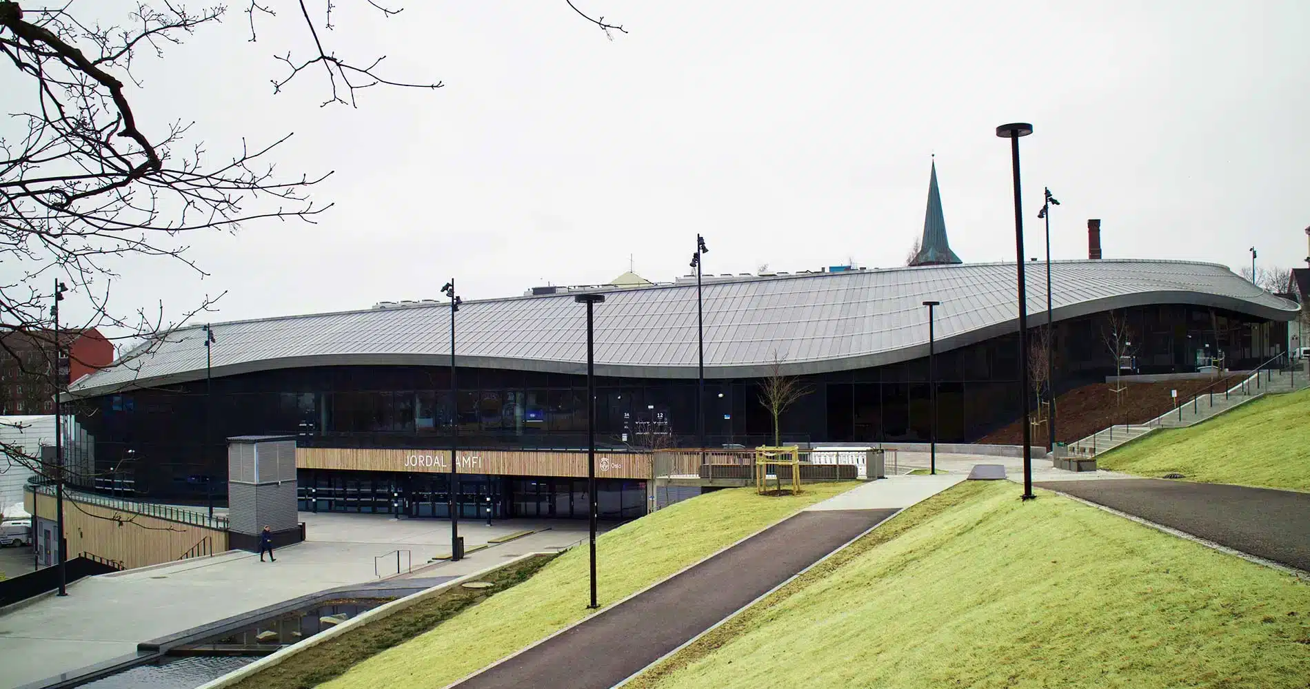 Bak en gressbakke og asfalterte gangveier ligger en moderne bygning med et bølgende, grått tak og store glassvinduer en overskyet dag.