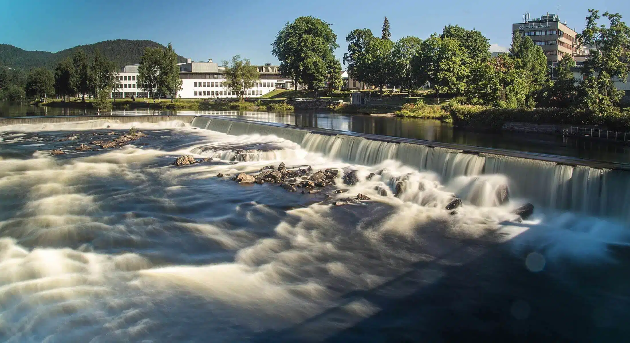 En elv med en lav foss renner forbi steiner og trær, med moderne bygninger synlige på motsatt bredd under en klarblå himmel.