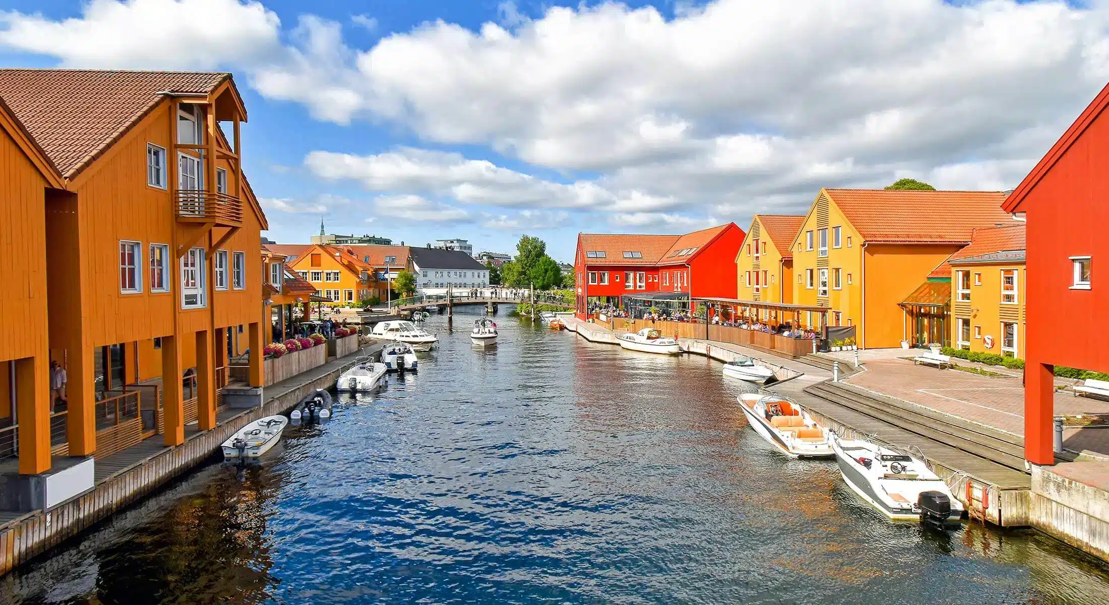 Fargerike trebygninger står på begge sider av en kanal med småbåter som ligger til kai langs kantene under en delvis overskyet himmel.