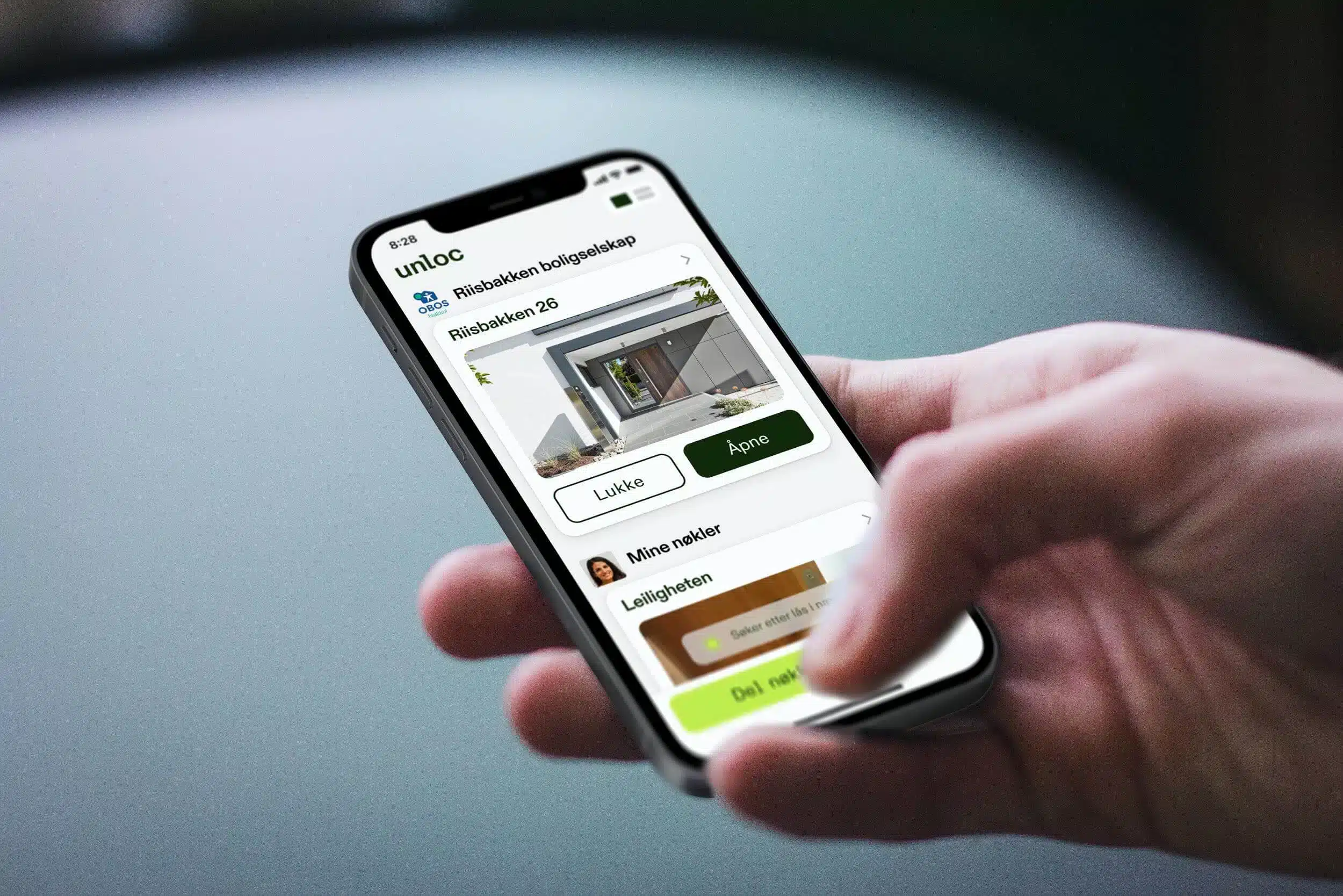 En hånd holder en smarttelefon som viser en app med muligheter for å åpne eller lukke en garasjeport på "Risbakken 25". Grensesnittet viser knapper merket "Lukk" og "Åpne".