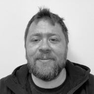 magne-3-1-e1642076295857 En mann med skjegg og bart, iført mørk hettegenser, står vendt mot kameraet og smiler lett mot en vanlig hvit bakgrunn.