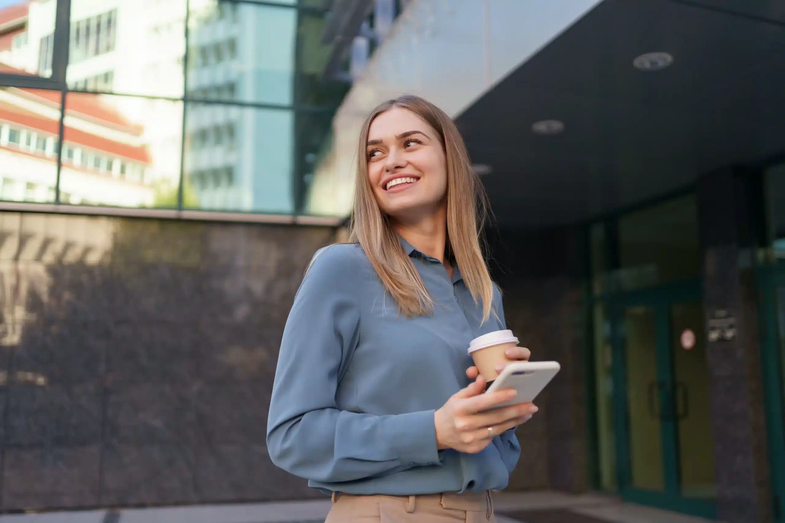 En kvinne står utendørs med en smarttelefon og en kaffekopp i hånden og smiler, med moderne glassbygninger i bakgrunnen.