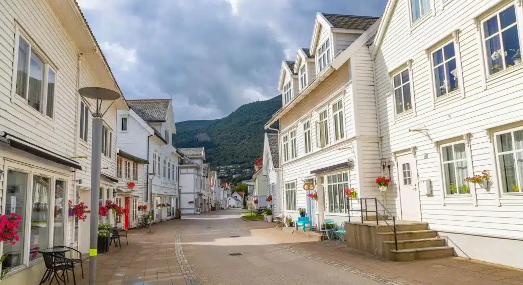En stille gate med hvite trehus og blomsterpotter, med fjellene synlige i bakgrunnen under en overskyet himmel.