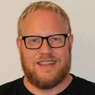 En mann med kort, blondt hår, skjegg og svarte briller, iført svart skjorte, smiler til kameraet mot en vanlig, lys bakgrunn.