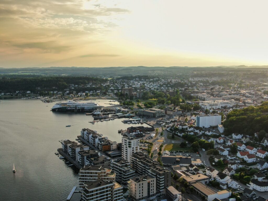 Luftfoto av en kystby med moderne bygninger, en havn med skip, boligområder og grønne åser under en disig himmel ved solnedgang.