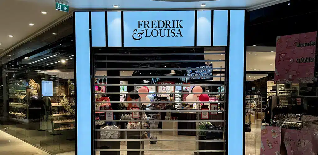 En Fredrik & Louisa-butikk med delvis lukkede sikkerhetsskodder av metall, plassert inne i et kjøpesenter med kosmetikk og produktutstillinger synlige på innsiden.