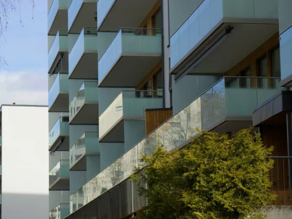 Moderne boligblokk med glassbalkonger og trepanel, sett fra en vinkel på en klar dag med et tre i forgrunnen.