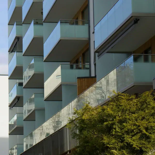Moderne boligblokk med glassbalkonger og trepanel, sett fra en vinkel på en klar dag med et tre i forgrunnen.