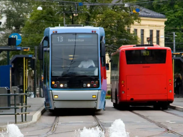 En blå trikk og en rød buss har stoppet på en bystasjon, med skinner og vannstråler i forgrunnen og bygninger i bakgrunnen.