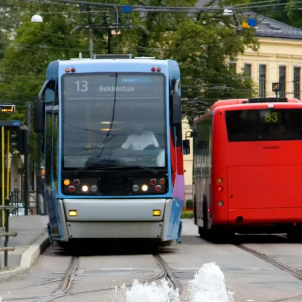 En blå trikk og en rød buss har stoppet på en bystasjon, med skinner og vannstråler i forgrunnen og bygninger i bakgrunnen.