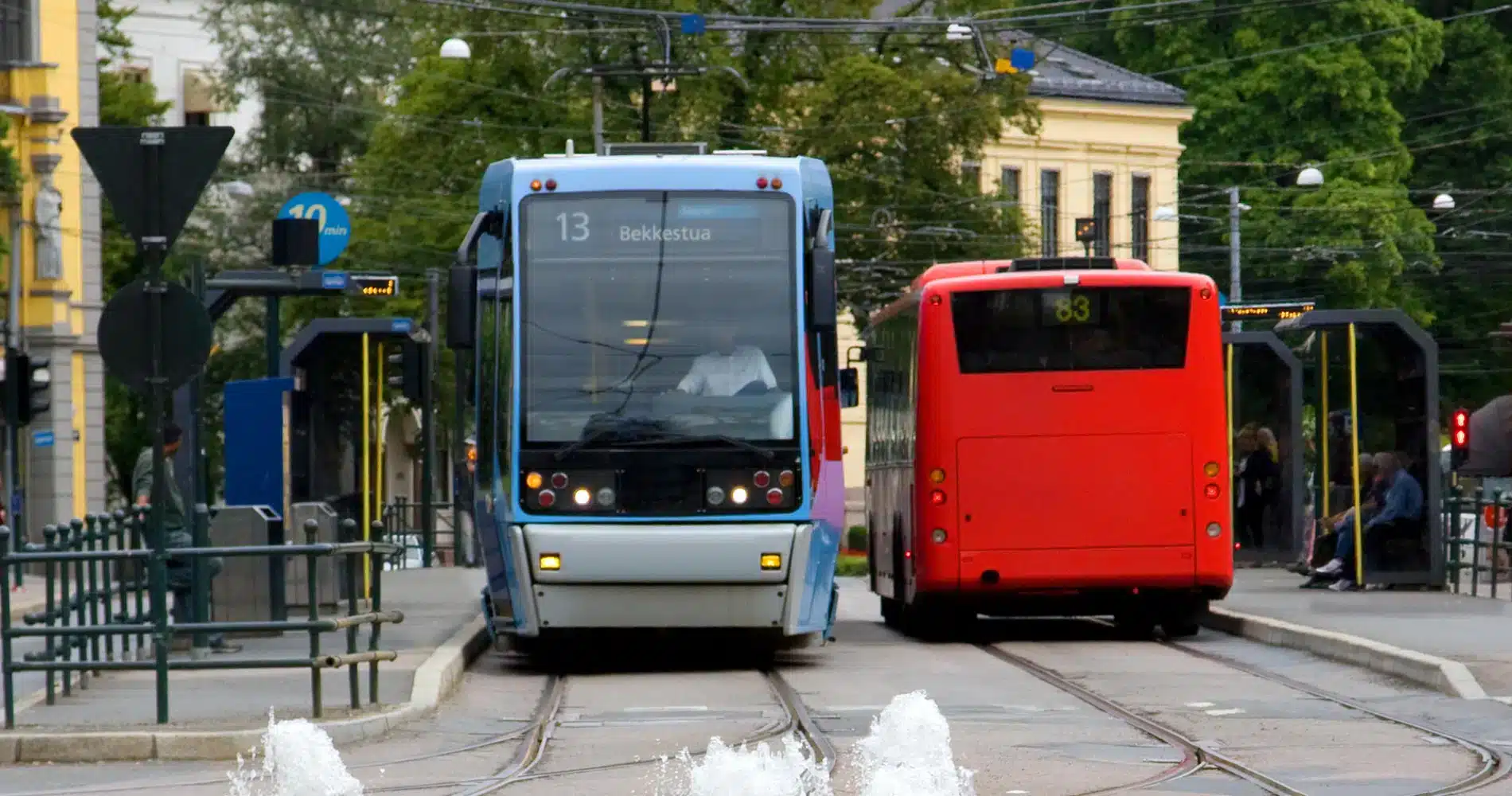 En blå trikk og en rød buss har stoppet på en bystasjon, med skinner og vannstråler i forgrunnen og bygninger i bakgrunnen.