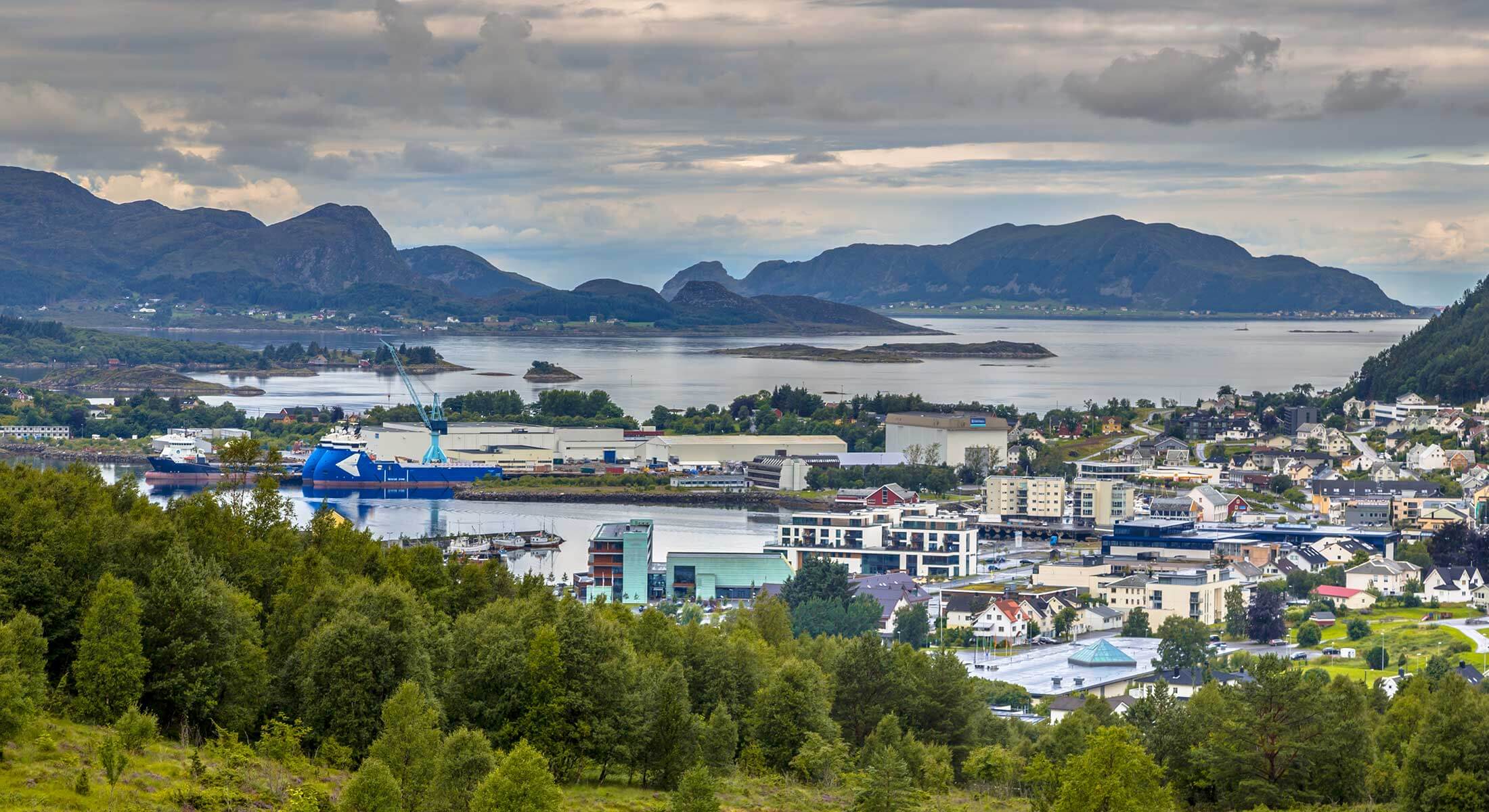 En kystby med industribygninger og kraner ligger ved en bukt, omgitt av grønne åser og fjell under en overskyet himmel.