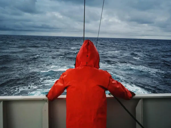 En person i oransje regnfrakk står ved rekka på en båt og ser ut over et overskyet, opprørt hav.