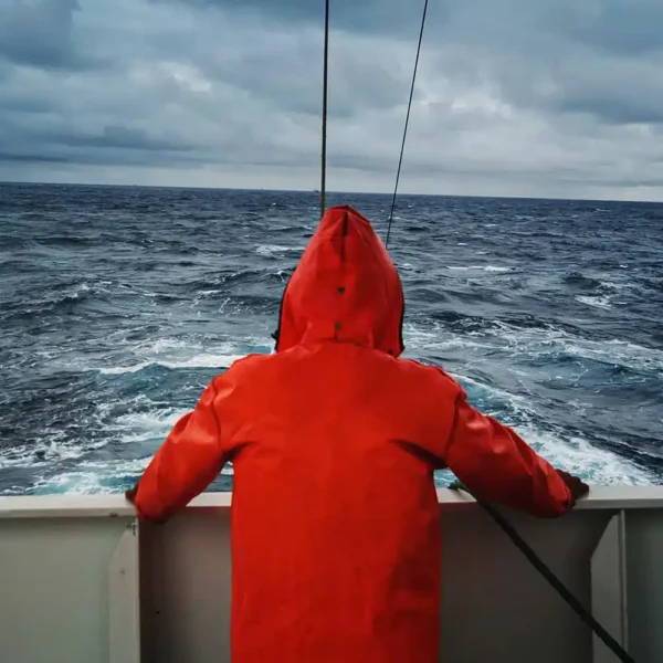 En person i oransje regnfrakk står ved rekka på en båt og ser ut over et overskyet, opprørt hav.