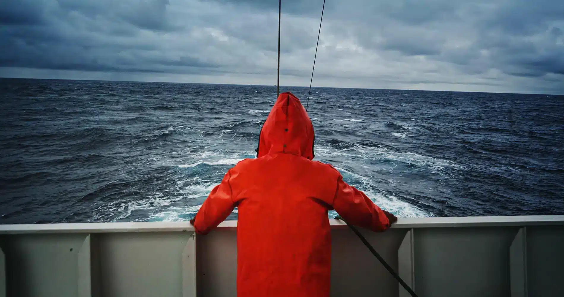 En person i oransje regnfrakk står ved rekka på en båt og ser ut over et overskyet, opprørt hav.