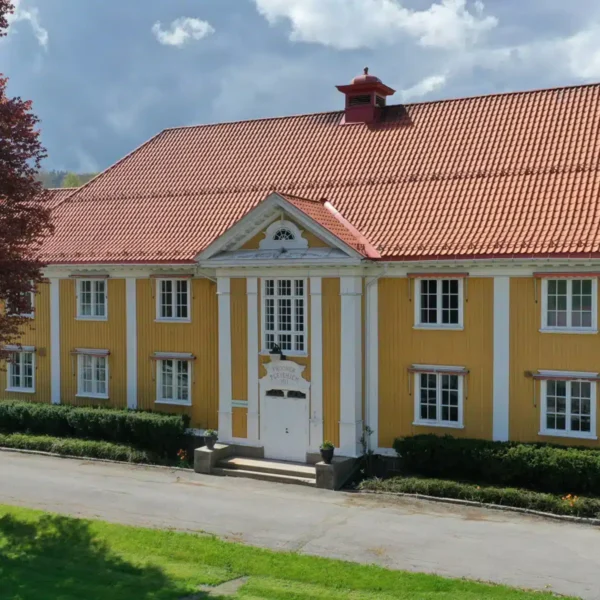 En stor, gul bygning i to etasjer med hvite detaljer, rødt tegltak, sentral inngang med søyler og flere vinduer, omgitt av grøntområder og et tre.