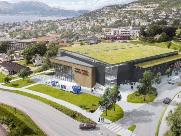 En stor, moderne bygning med grønt tak, busser parkert utenfor og folk som går i nærheten, i en kupert by med hus, trær og et vann i bakgrunnen.