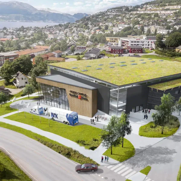 En stor, moderne bygning med grønt tak, busser parkert utenfor og folk som går i nærheten, i en kupert by med hus, trær og et vann i bakgrunnen.