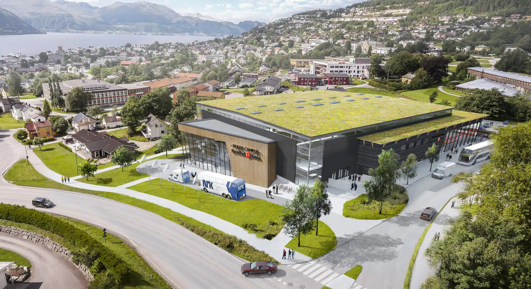 En stor, moderne bygning med grønt tak, busser parkert utenfor og folk som går i nærheten, i en kupert by med hus, trær og et vann i bakgrunnen.
