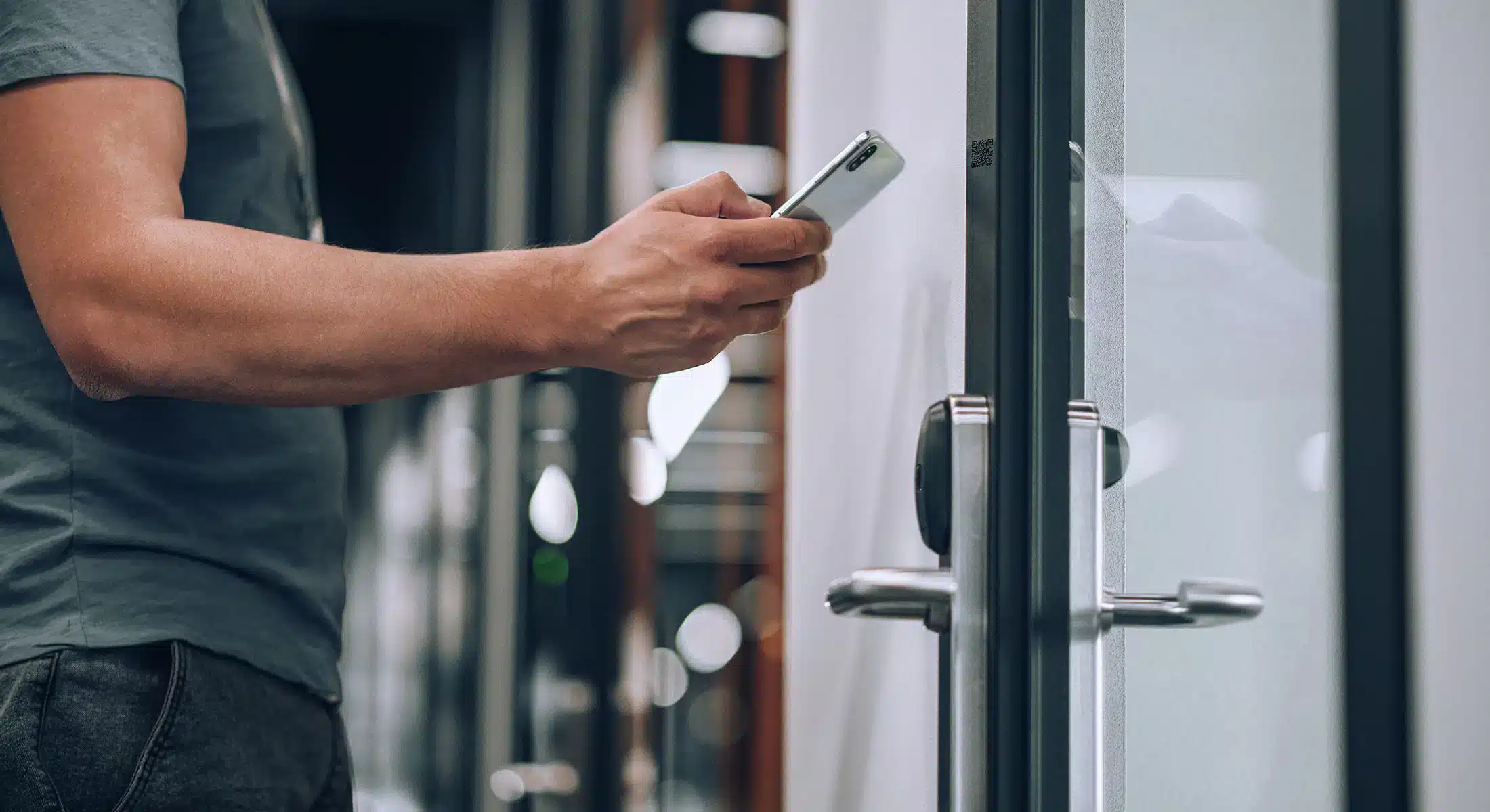 En person med smarttelefon står ved siden av en glassdør med elektronisk lås og ser ut til å låse den opp ved hjelp av enheten sin.